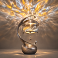 ORVAITOLEY® Art Meets Light – full product view on grey background showing dramatic golden flame projection across wall and ceiling, spiral silver crescent arm, artistic glass globe shade with gold swirl interior, metal dome cap, water-drop pendants and round stone-texture base